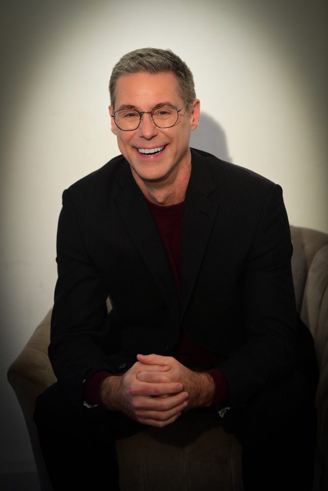 Roy Samuelson — a wide grin of recognition, wearing glasses and a dark suit, seated with a spotlight shadow on the wall behind him