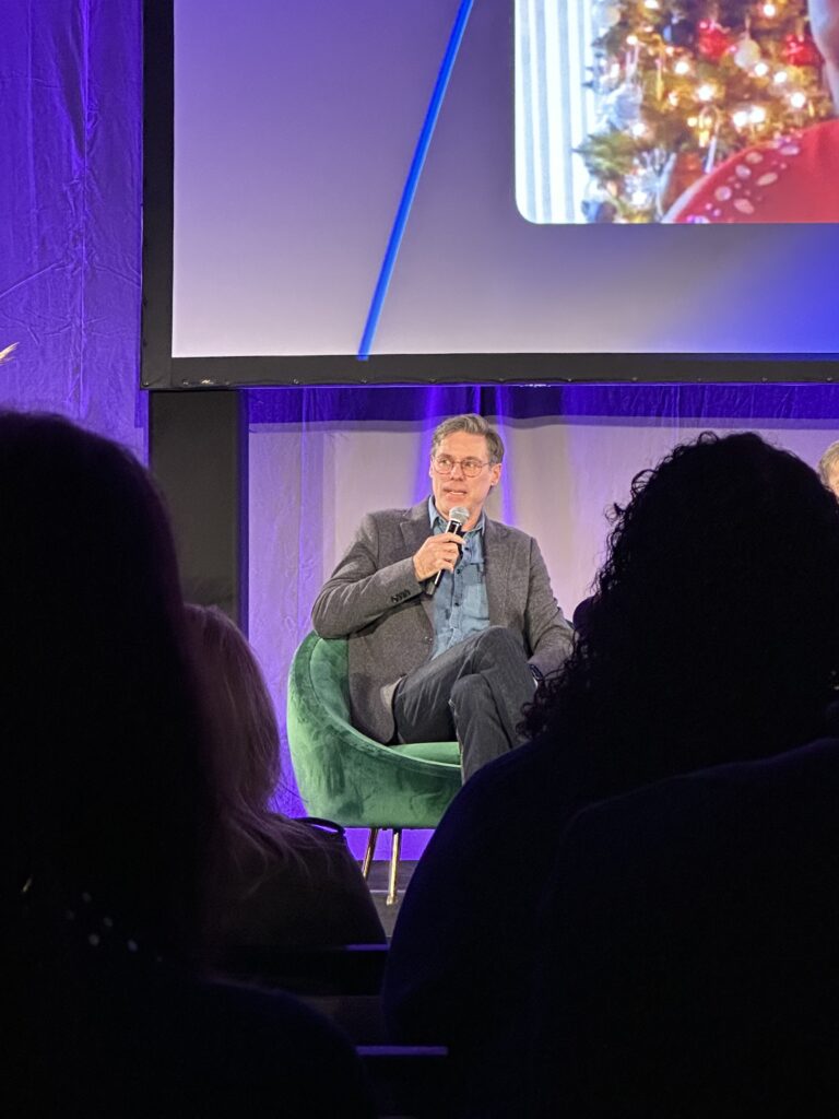 Seen between silhouetted audience members, Roy sits with crossed legs, beneath a large screen. He wears glasses and a soft gray suit jacket, and holds a microphone.